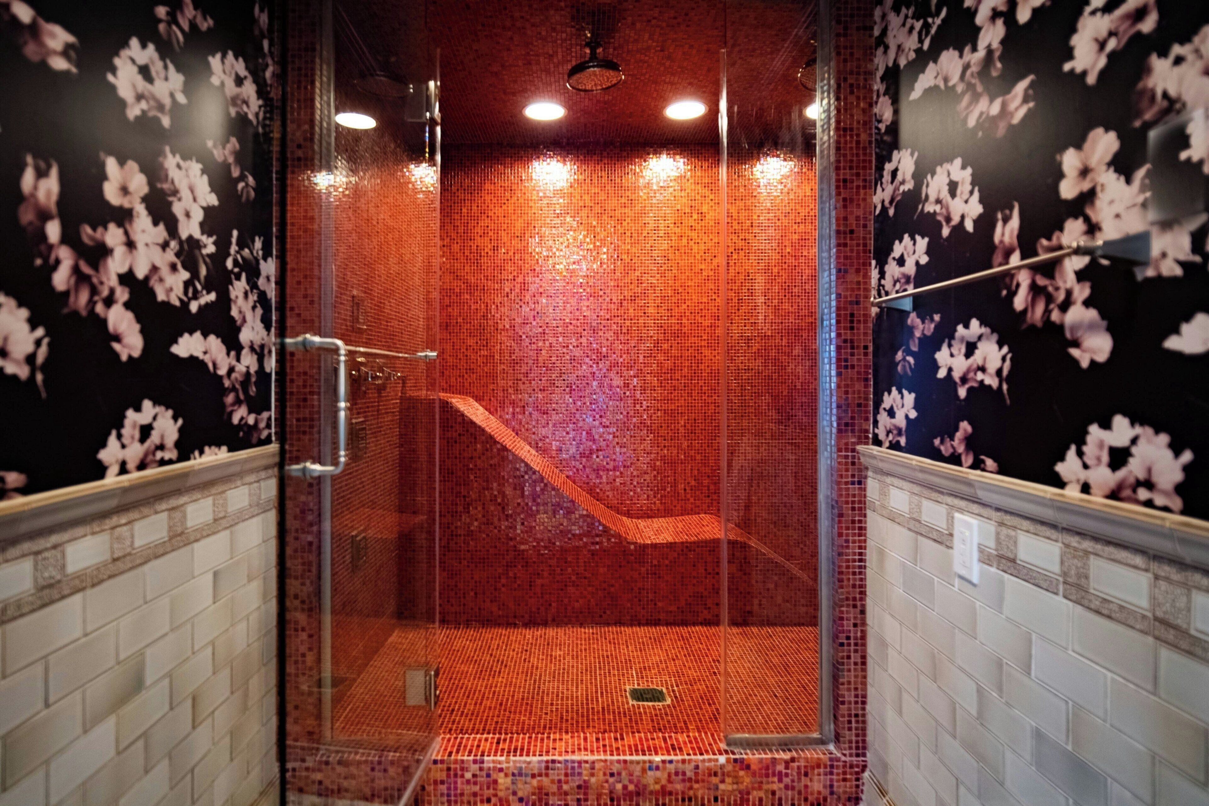 Une douche fermée par des parois en verre, avec un sol et des murs carrelés, une pomme de douche en métal et un drain visible.