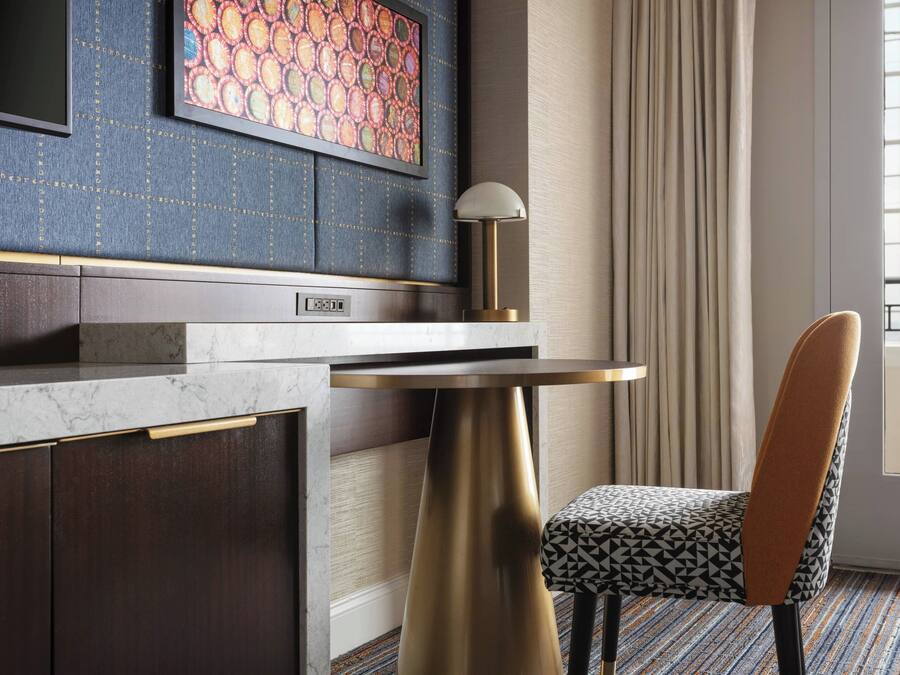 A modern hotel room with a marble countertop, a gold lamp, a patterned chair, and a large window with curtains.