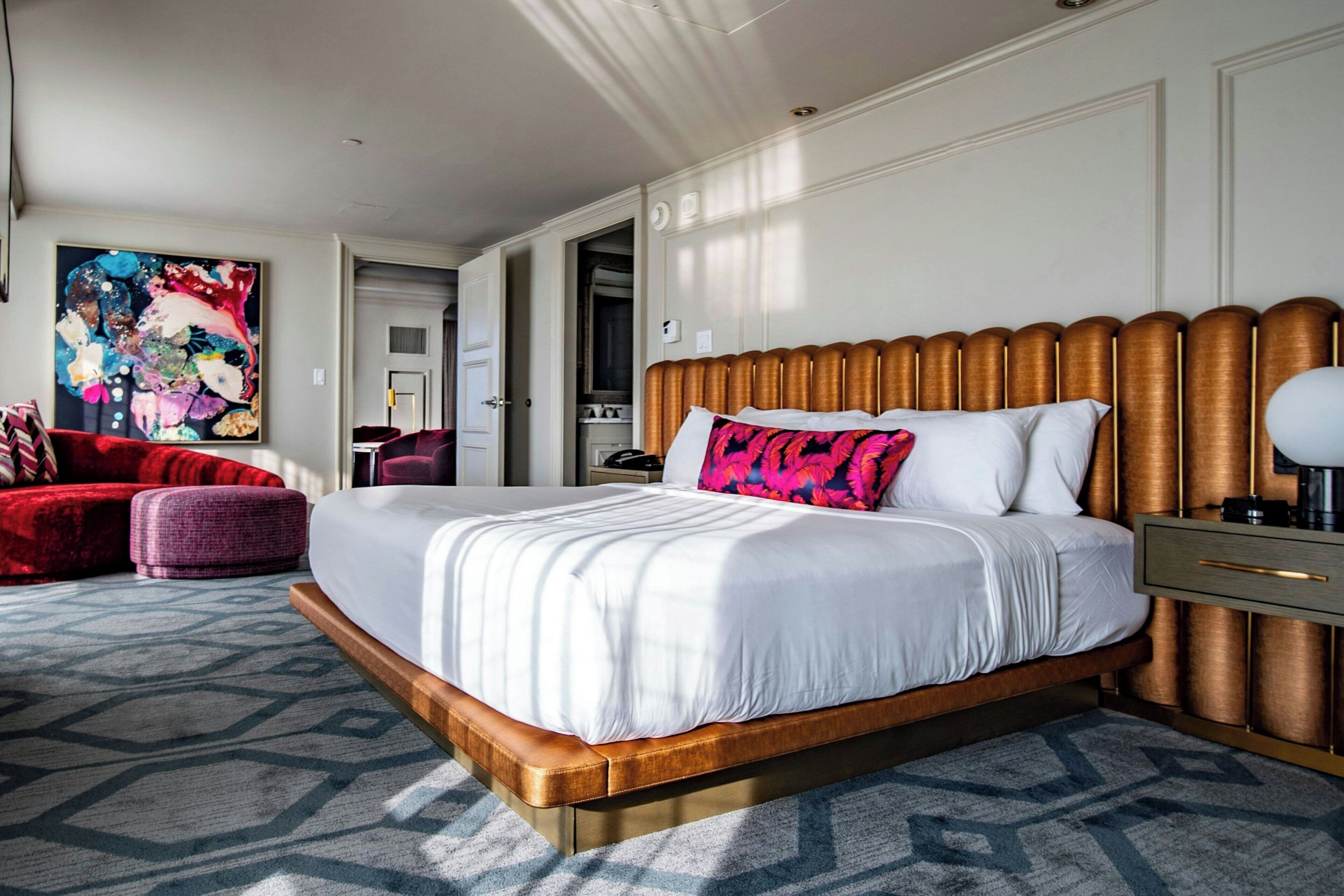 A hotel room with a large bed, a red sofa, a purple ottoman, and a colorful abstract painting on the wall.
