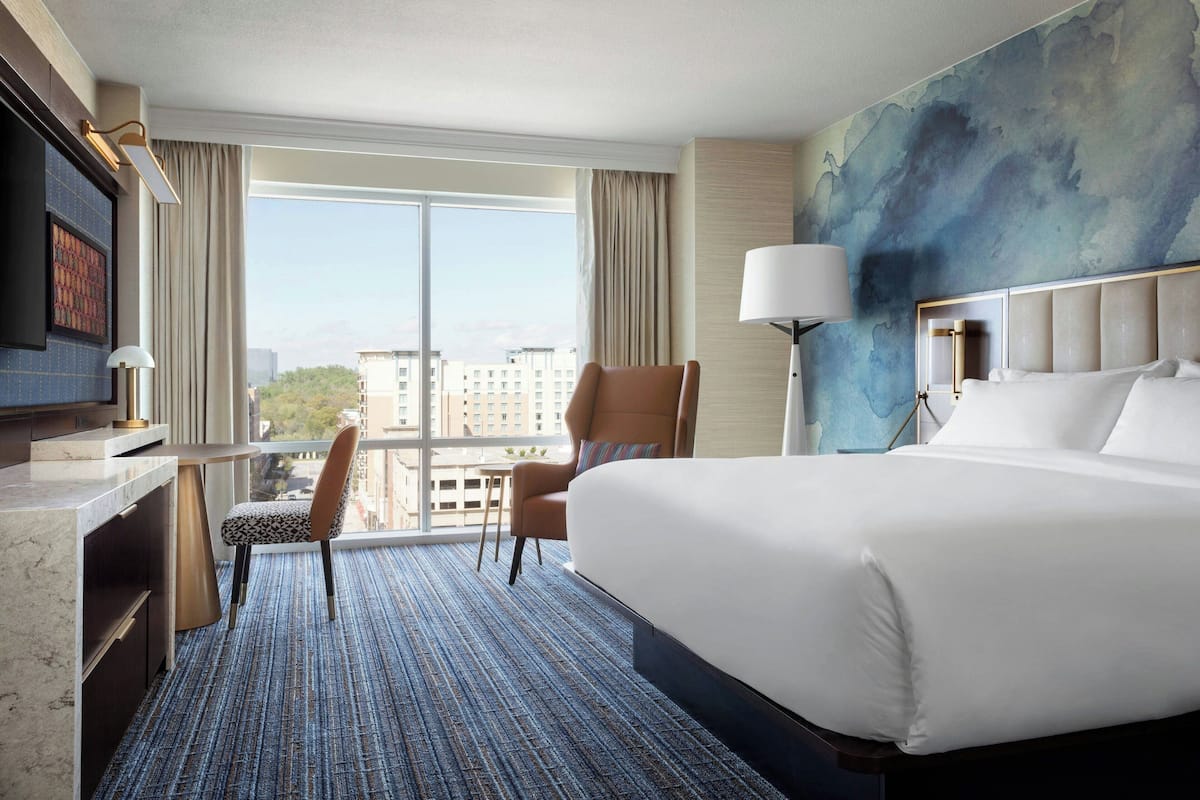Hotelkamer met een groot bed, een bureau met stoel, een televisie en uitzicht op de stad door grote ramen.