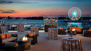 Un espace extérieur aménagé au bord de l’eau, avec des foyers, des meubles en osier et une vue sur une grande roue illuminée.