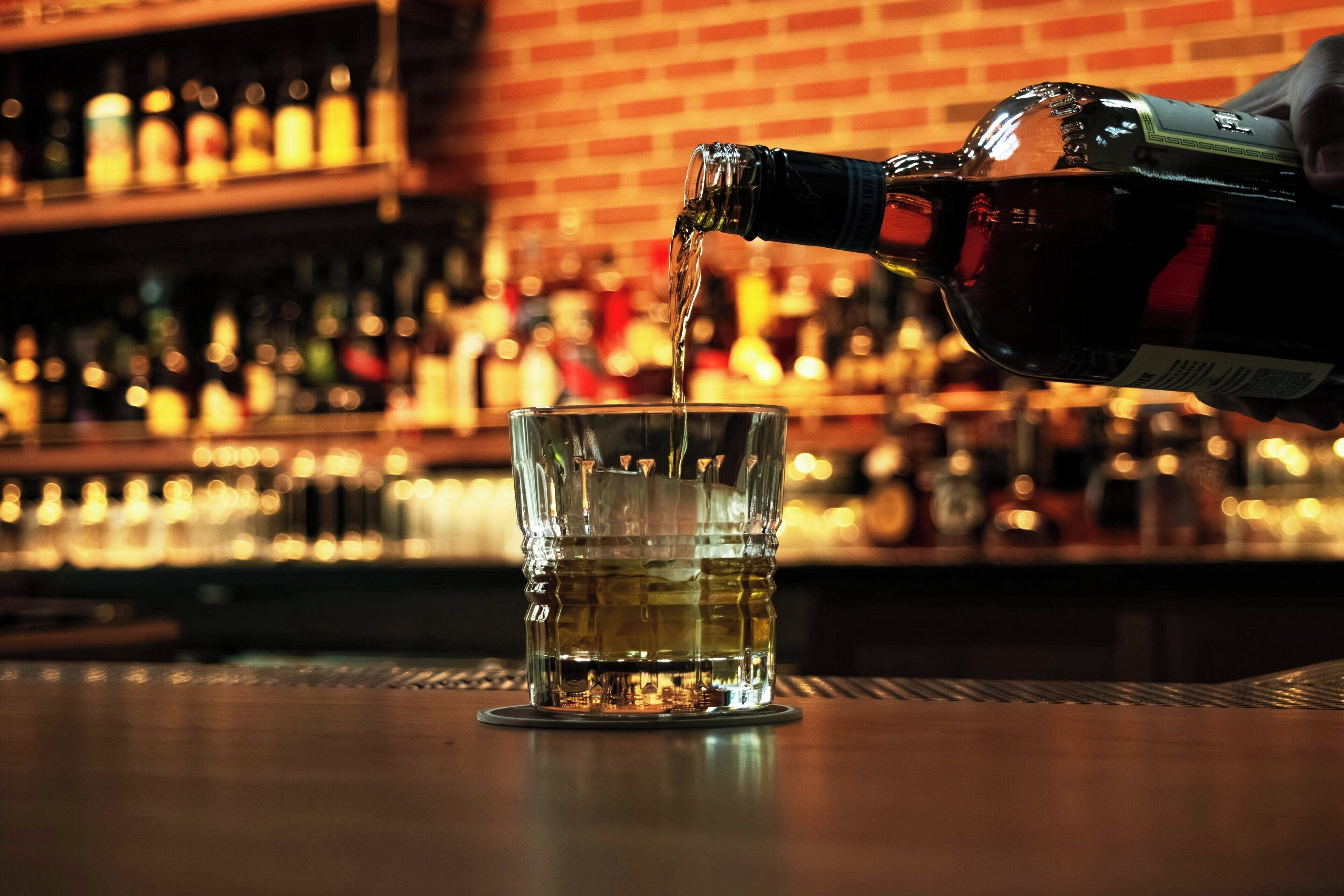 A bar setting with a glass being filled with a dark liquid, likely whiskey, and a variety of bottles in the background.