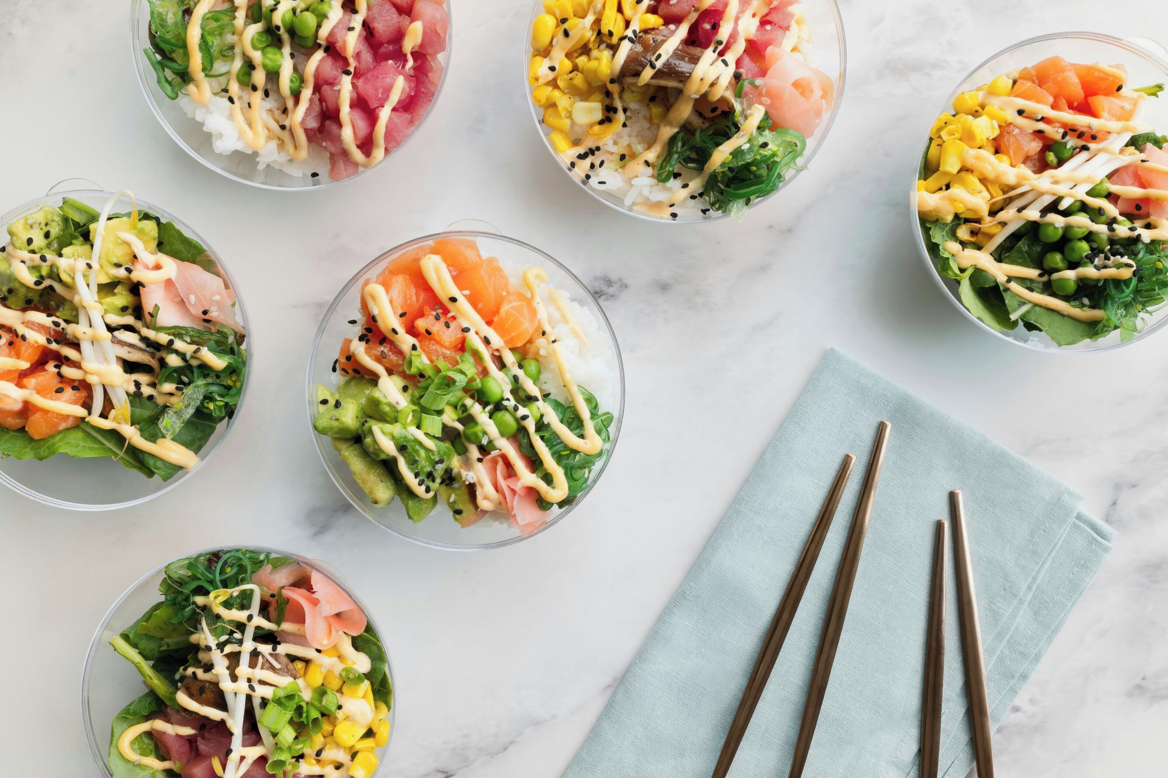 Six bowls of colorful salad with various toppings and sauces, arranged on a marble surface with chopsticks and a napkin.