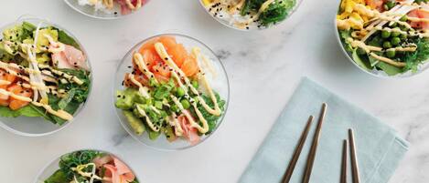 Six bowls of colorful salad with various toppings and sauces, arranged on a marble surface with chopsticks and a napkin.