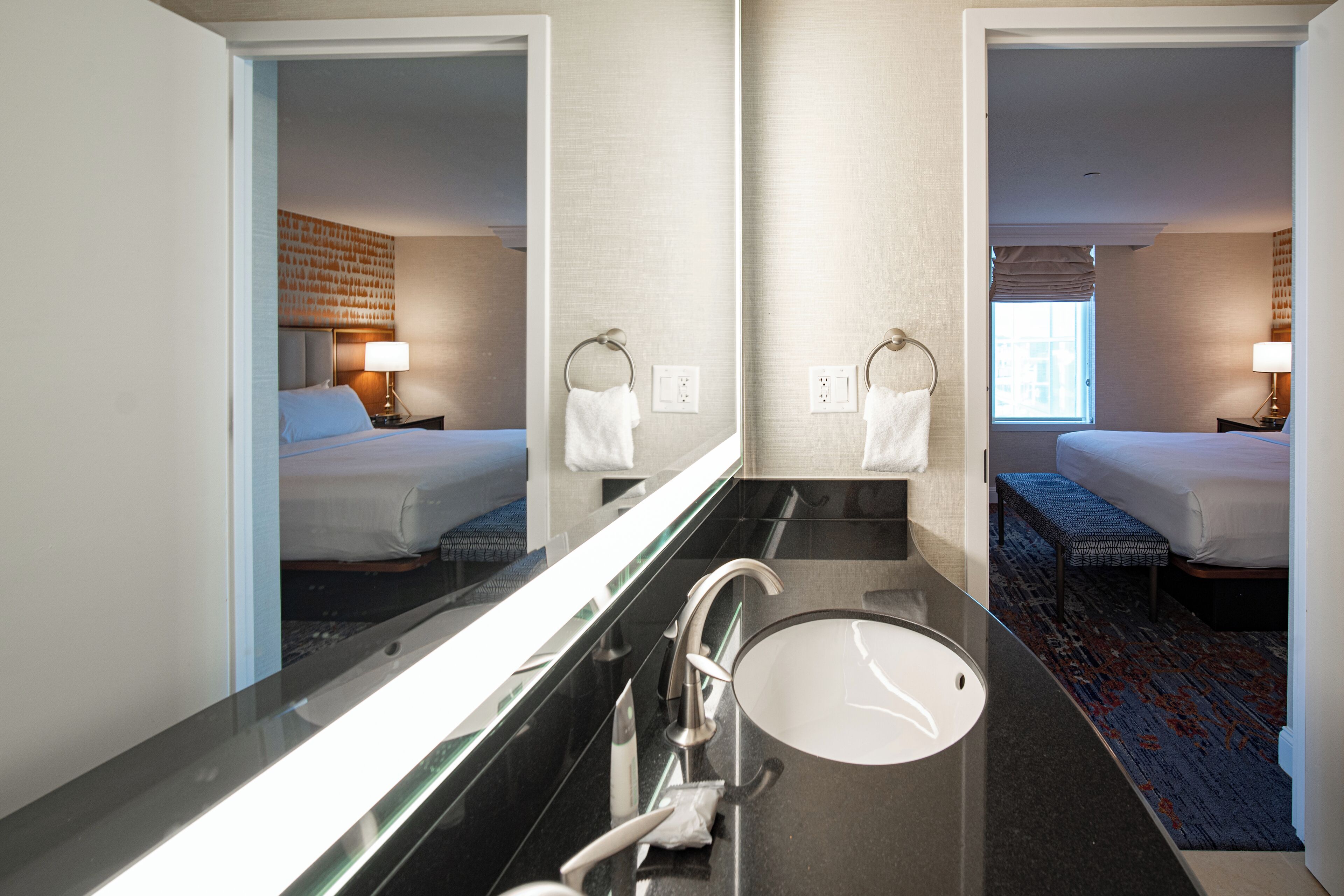 Un baño de hotel con un espejo grande, un lavamanos y una cama visible en el reflejo.