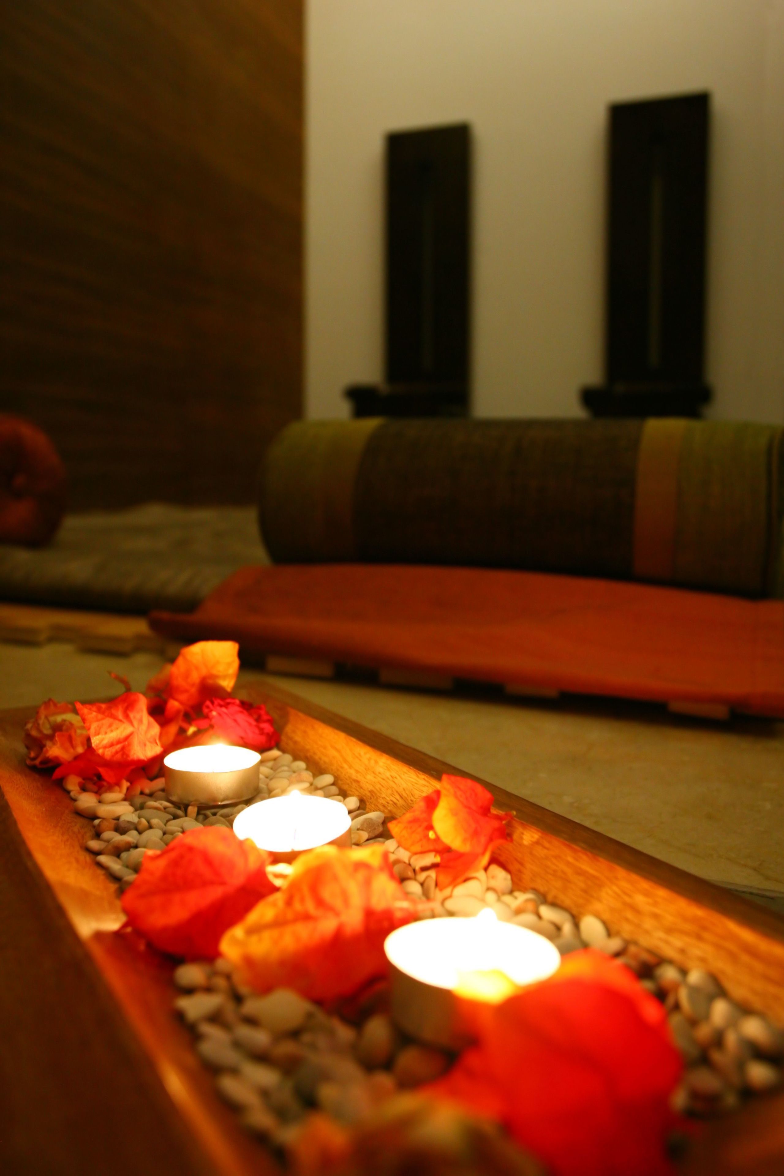 A candle-lit pathway with red flowers and pebbles, leading to a seating area with a green cushion and a wooden bench.