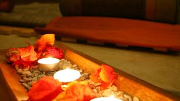A candle-lit pathway with red flowers and pebbles, leading to a seating area with a green cushion and a wooden bench.