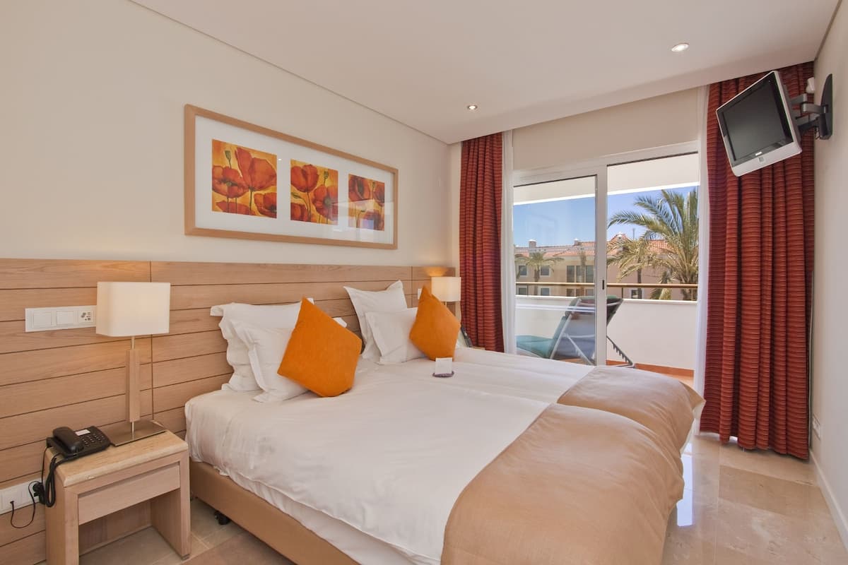A hotel room with a large bed, a bedside table with a phone, a wall-mounted TV, and a balcony with red curtains.