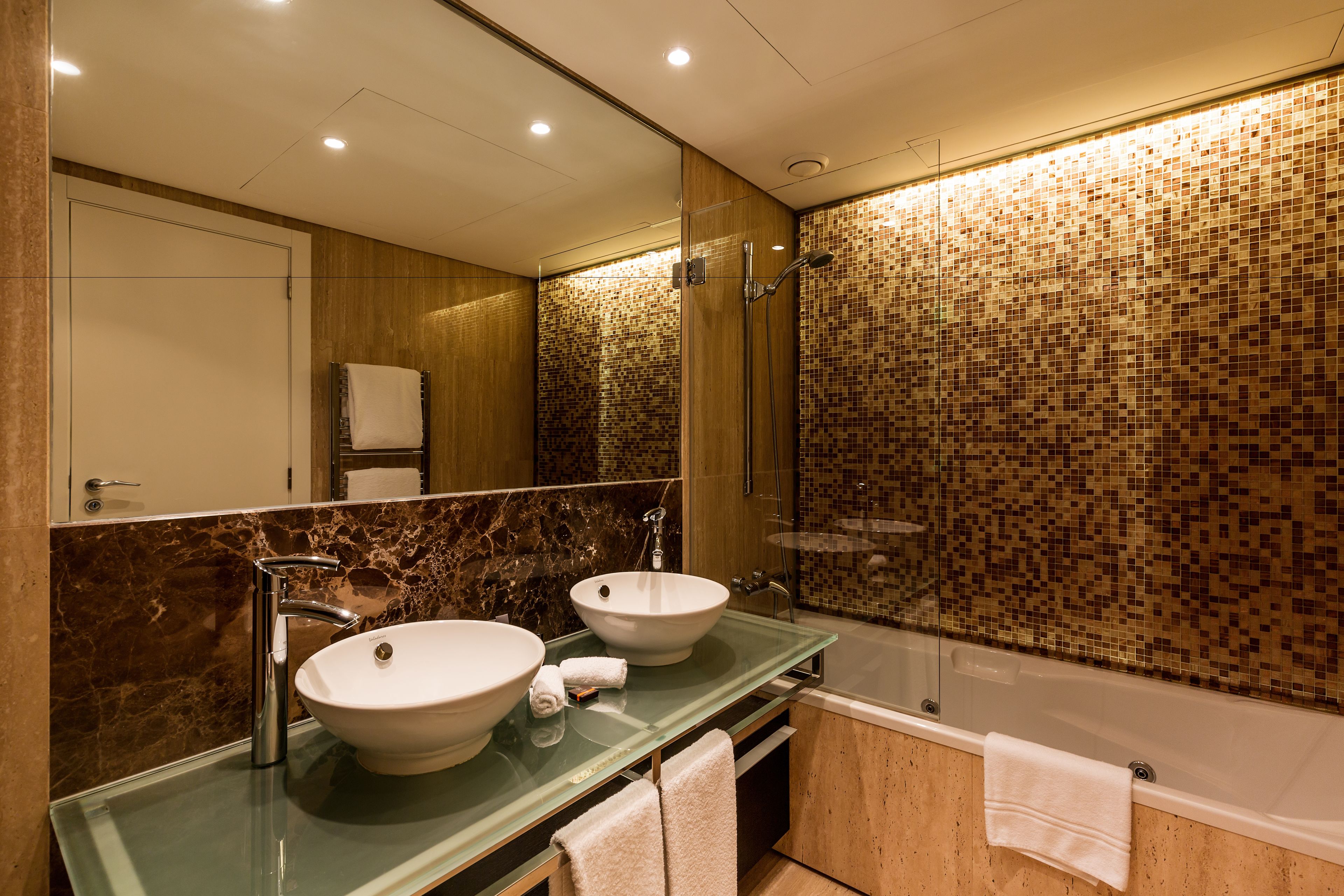 Une salle de bain moderne avec deux vasques, un meuble-lavabo en verre, un grand miroir et un mur de douche recouvert de mosaïques.