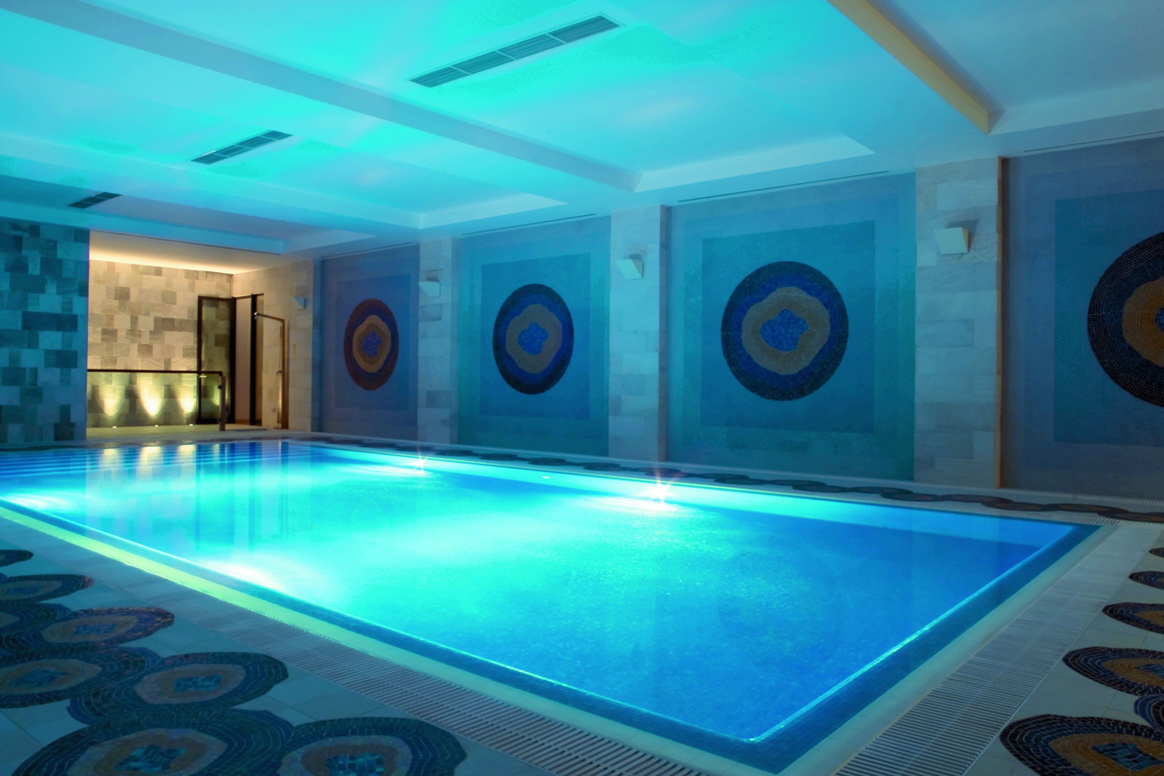 A large indoor swimming pool with a blue-lit edge, surrounded by patterned tiles and circular wall art.