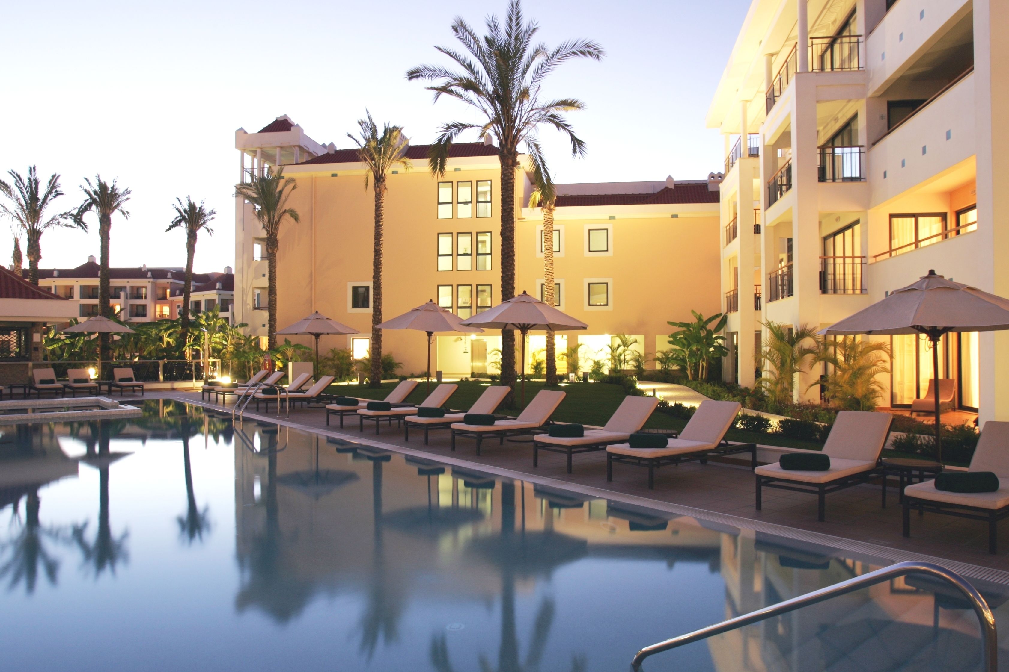 A pool area with lounge chairs and umbrellas, surrounded by palm trees and a hotel building.