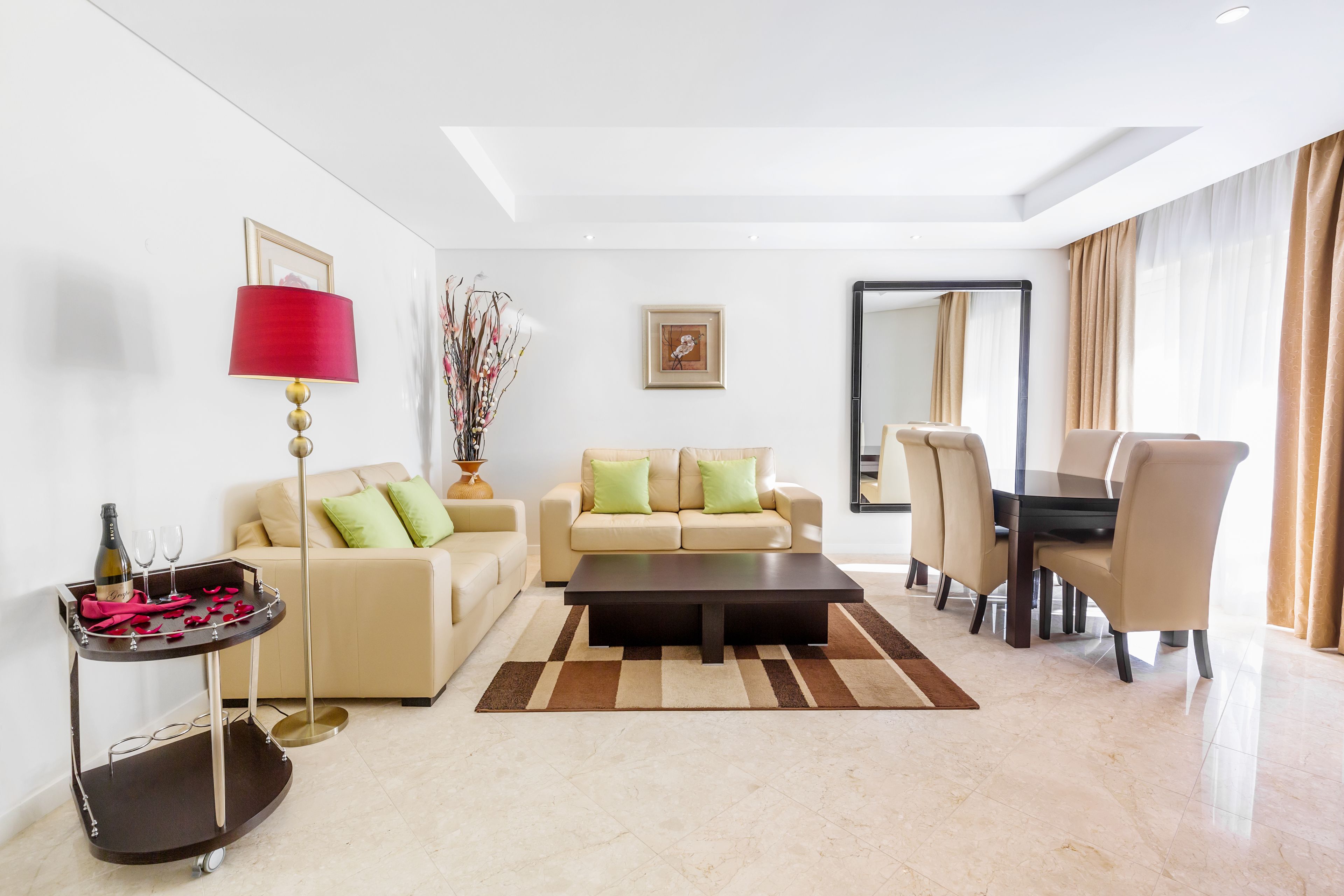 Uma sala de estar moderna com um sofá bege, uma mesa de centro, uma mesa de jantar com cadeiras, um abajur vermelho e um vaso decorativo.
