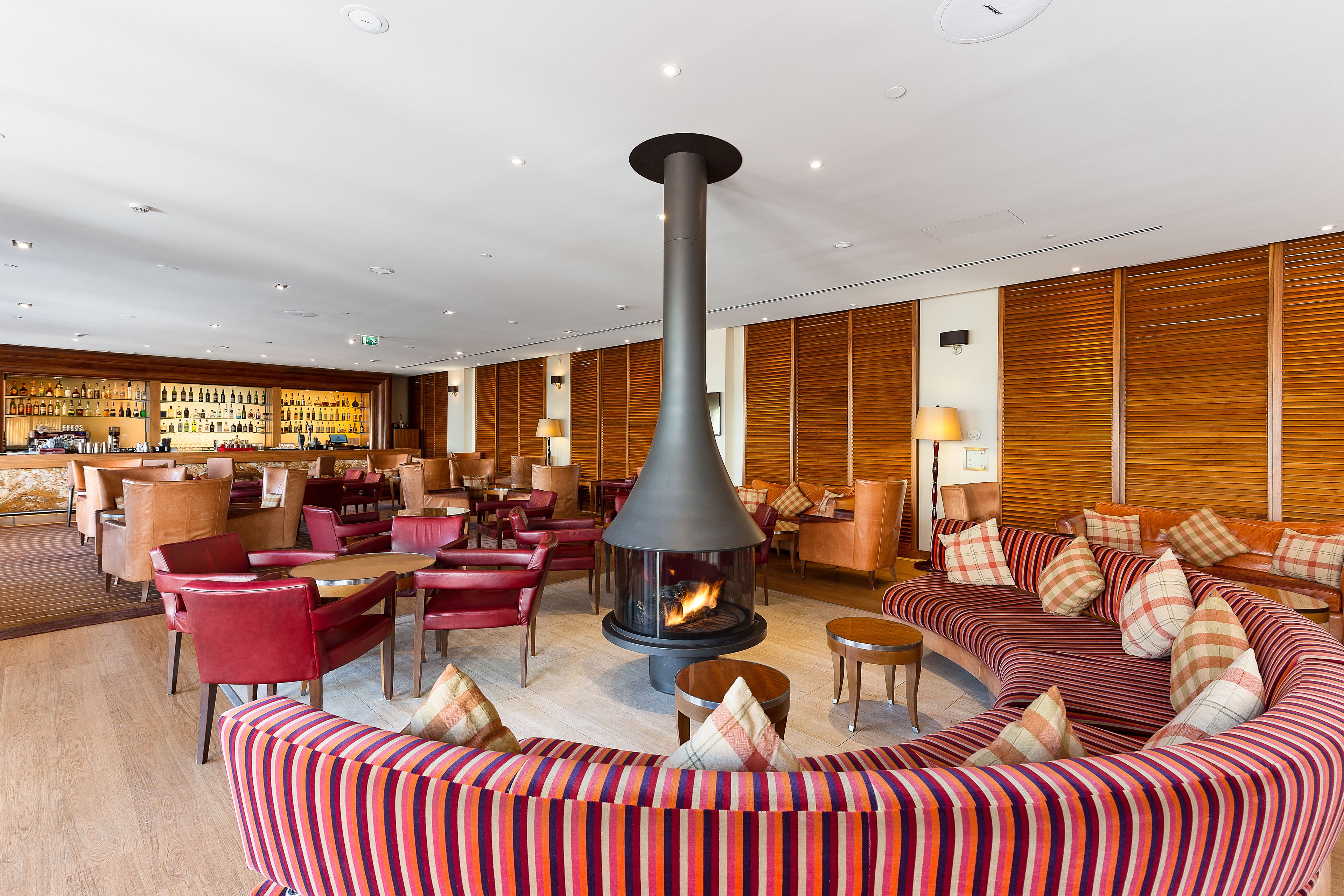 A modern lounge area with a central fireplace, red upholstered seating, and a bar in the background.