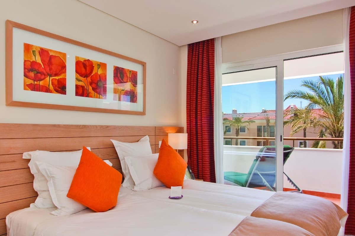 A hotel room with a large bed, red curtains, a balcony with a view of palm trees and buildings, and framed artwork on the wall.