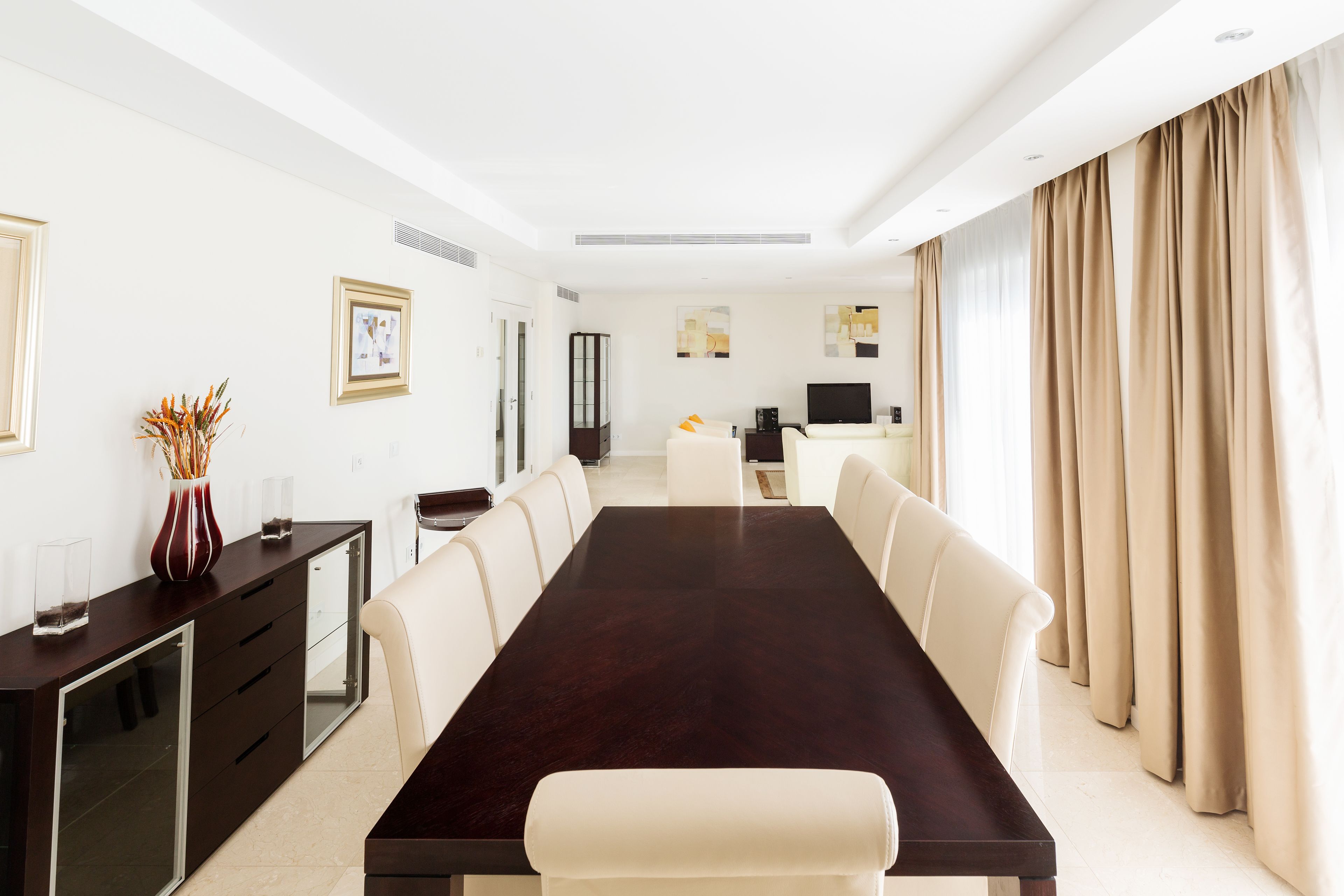 A modern hotel room with a large dark wood dining table, white leather chairs, a minibar, a flat-screen TV, and beige curtains.