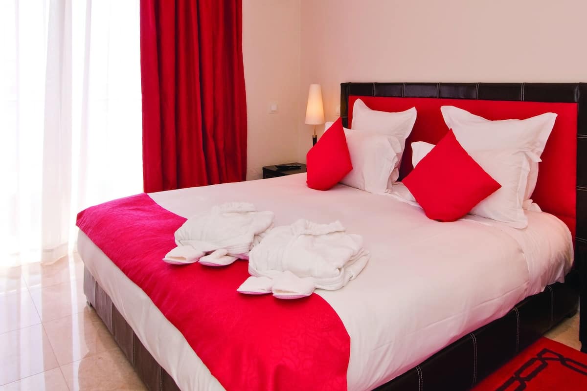 A hotel room with a large bed, red and white bedding, a red headboard, and a nightstand with a lamp.