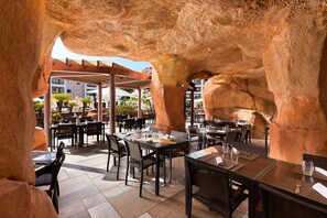 An outdoor dining area with tables and chairs, a rock arch structure, and a view of buildings in the background. - As Cascatas Golf Resort & Spa (Loulé)