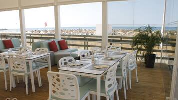 A restaurant with white furniture, a wooden floor, and a view of the beach.