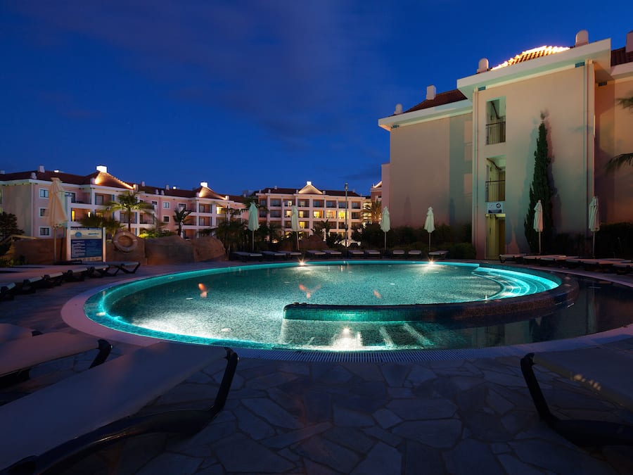 A swimming pool with a fountain, surrounded by lounge chairs and palm trees, with hotel buildings in the background.