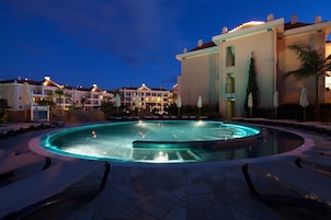 A swimming pool with a fountain, surrounded by lounge chairs and palm trees, with hotel buildings in the background.