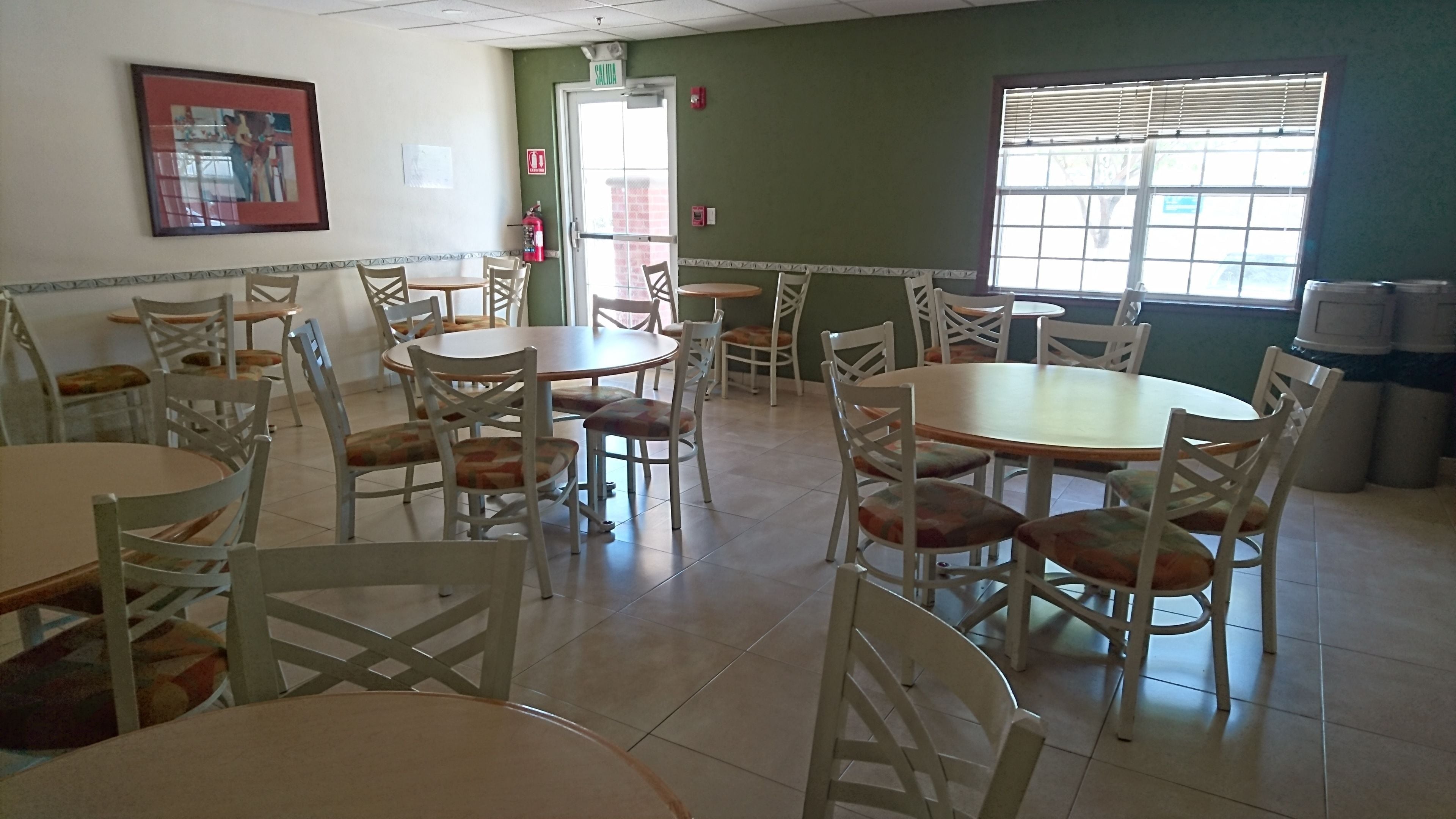 A dining area with multiple round tables and chairs, a fire extinguisher, a door with a 'SALIDA' sign, and a window with blinds.