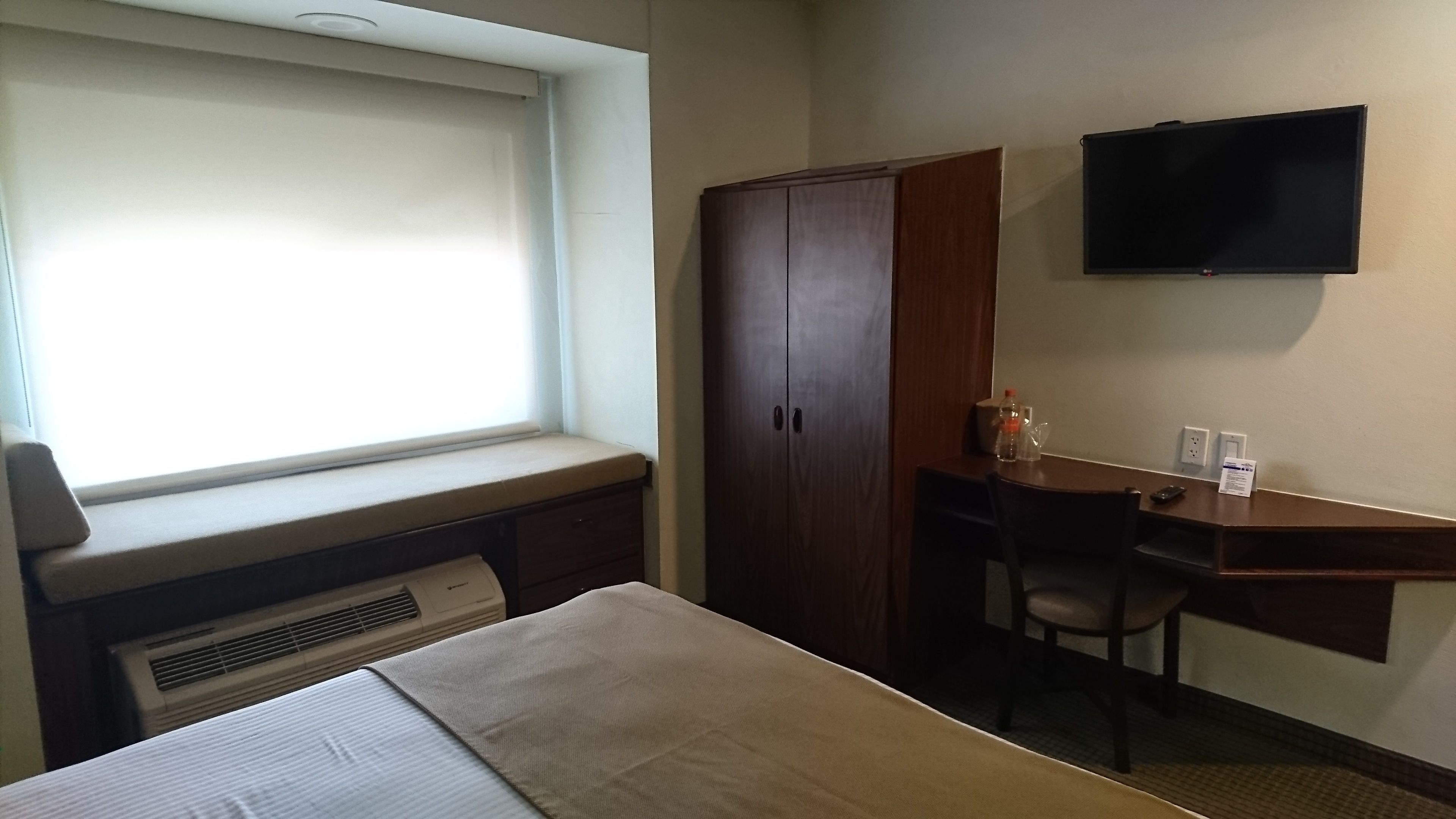 Une chambre d’hôtel comprenant un lit, un bureau avec une chaise, une armoire, un téléviseur fixé au mur et une fenêtre avec des stores.