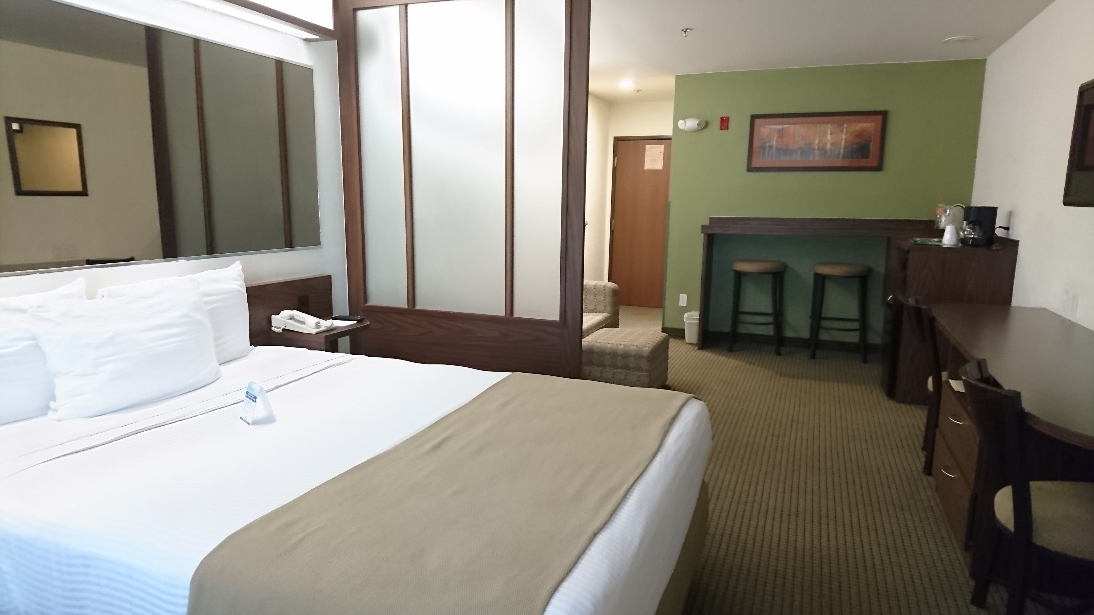 A hotel room with a large bed, a bedside telephone, a small table, a desk with two stools, and a green wall with a painting.