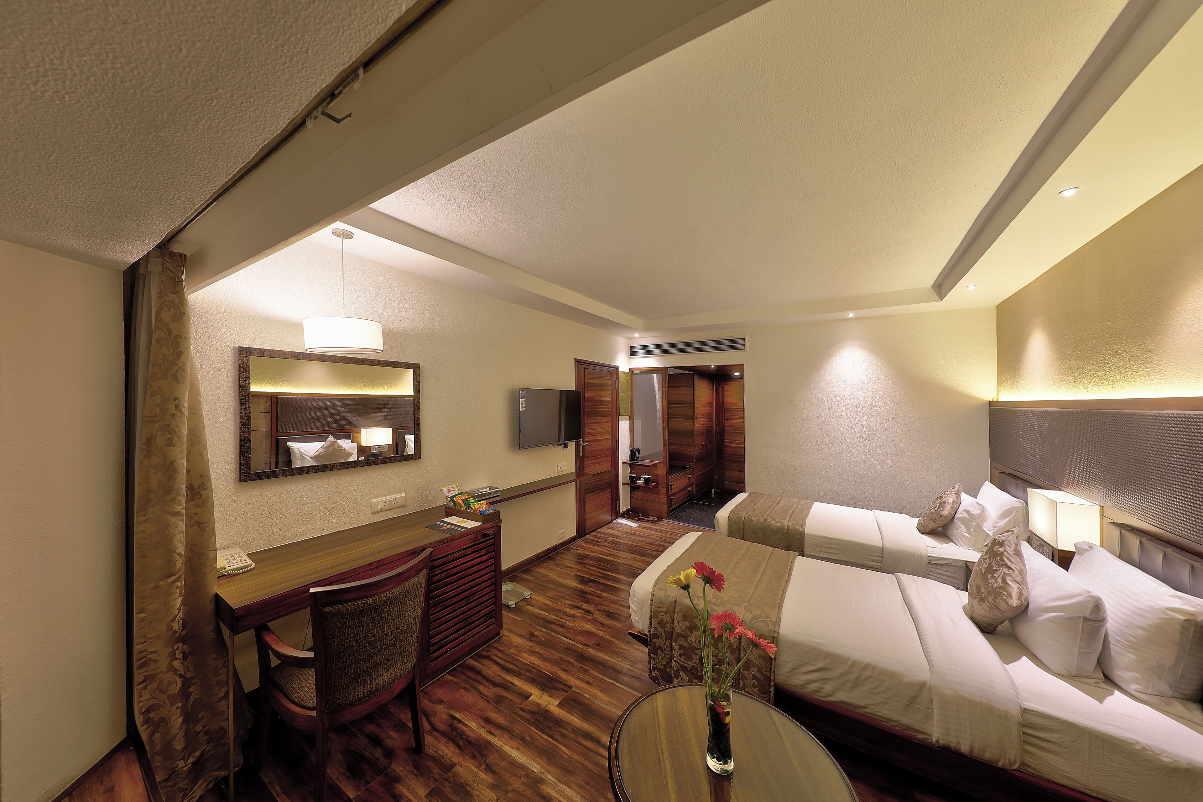 Habitación de hotel con escritorio, dos camas, espejo, televisor y puerta de baño.