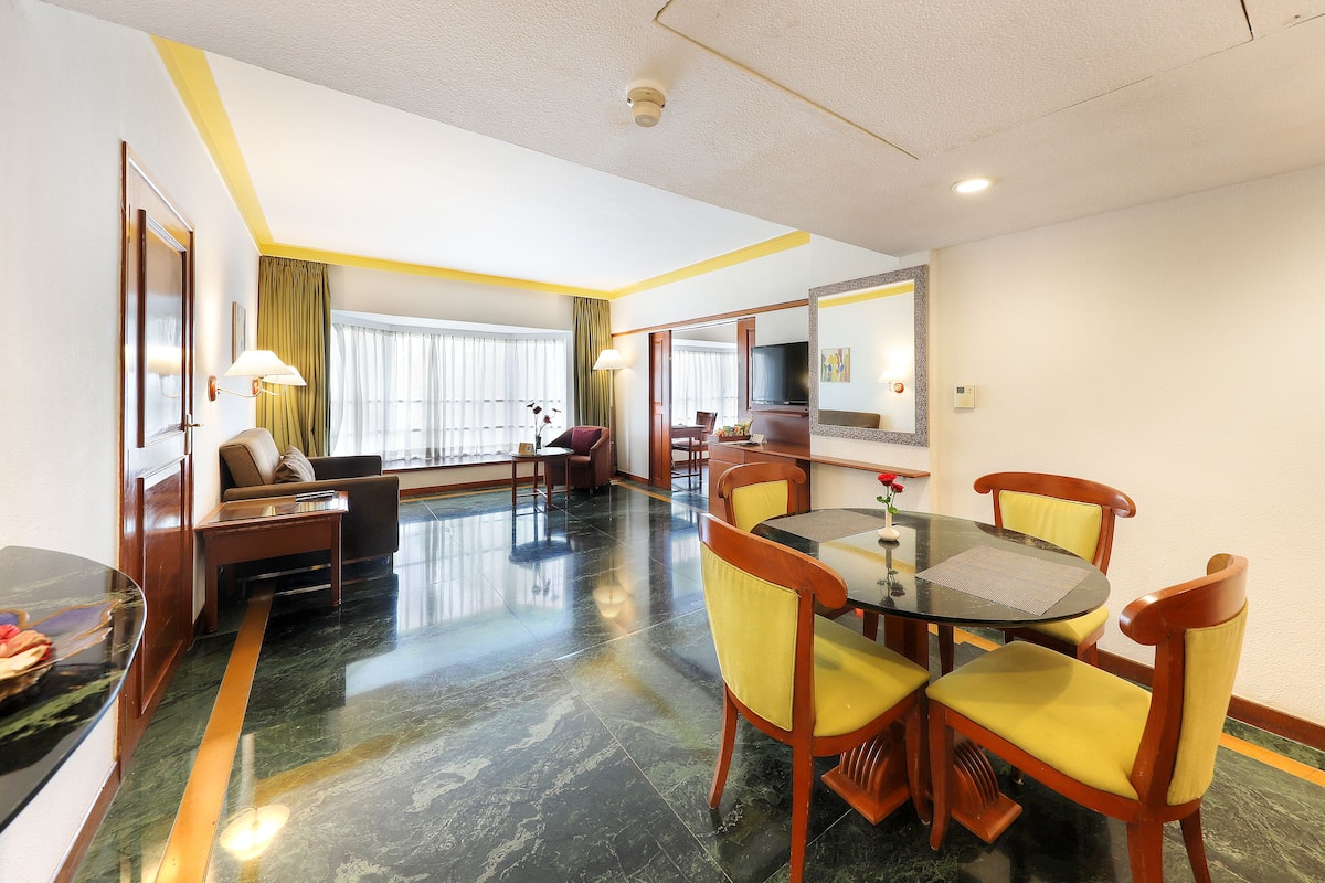 A hotel room with a dining area, a sofa, a TV, and a large window with curtains.