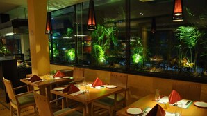 Un restaurante con mesas y sillas de madera, un amplio ventanal con plantas tropicales e iluminación colgante.