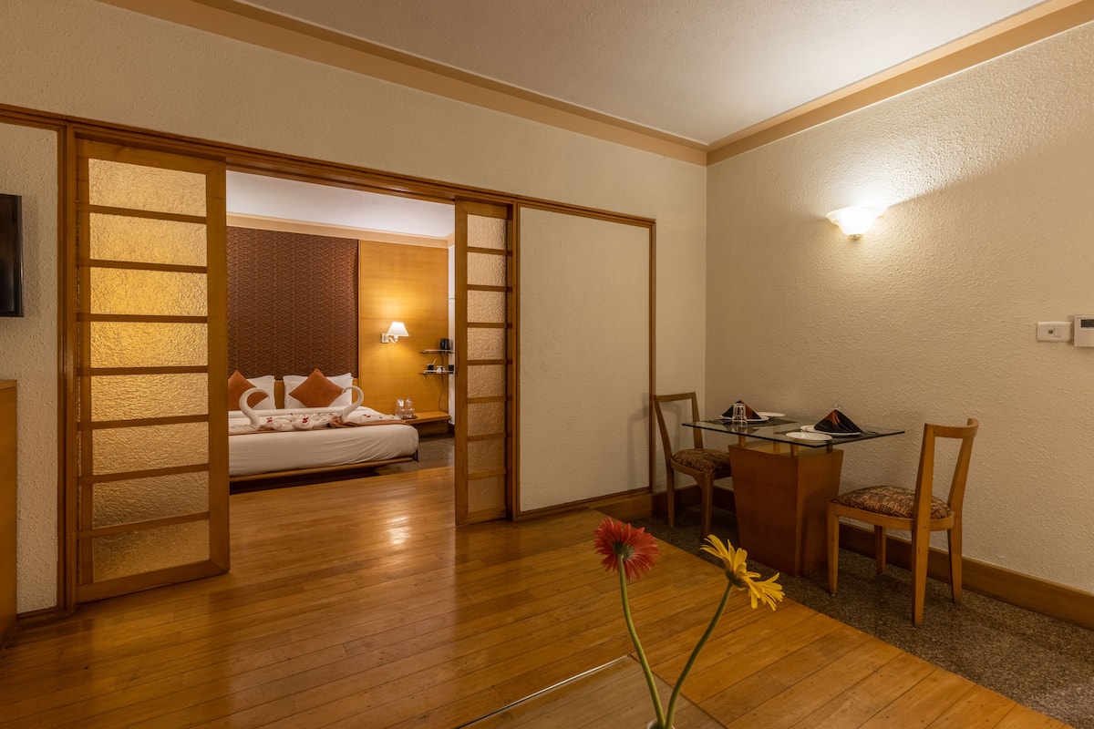 A hotel room with a wooden floor, a bed with pillows, a desk with a chair, and a vase with flowers.