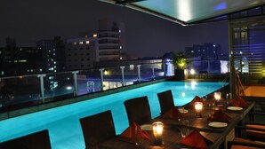 Un área de piscina en la azotea con mesas y sillas, vistas a la ciudad y una zona de estar cubierta.