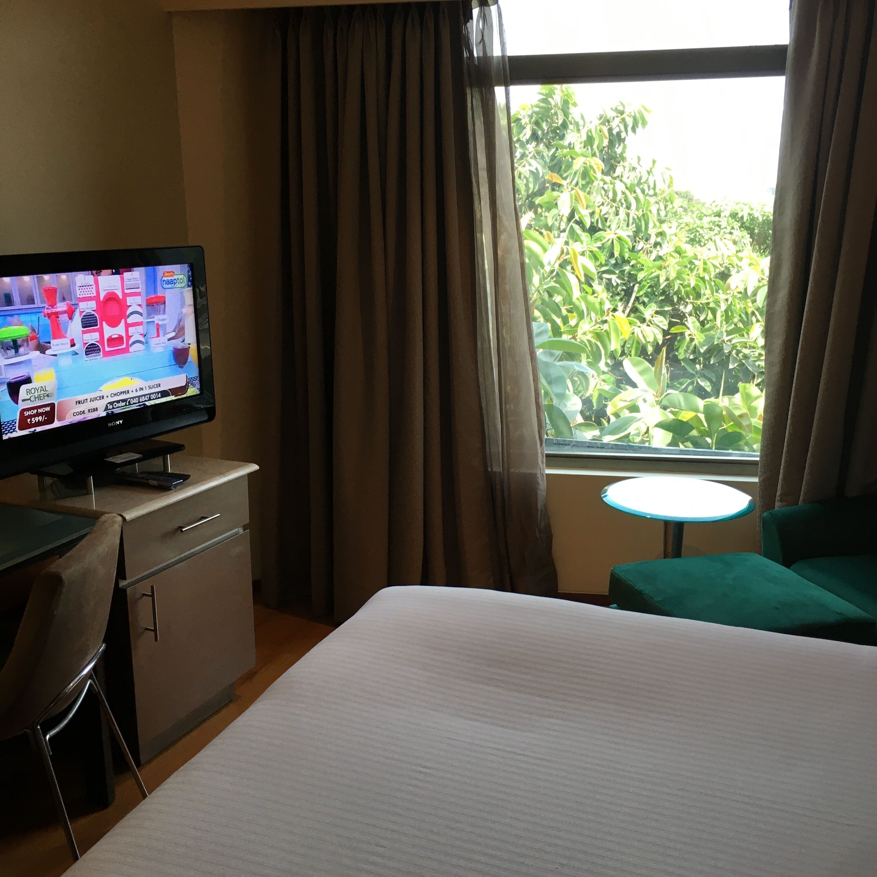 A hotel room with a bed, a desk with a television displaying a cooking show, a small round table, and a view of greenery outside.