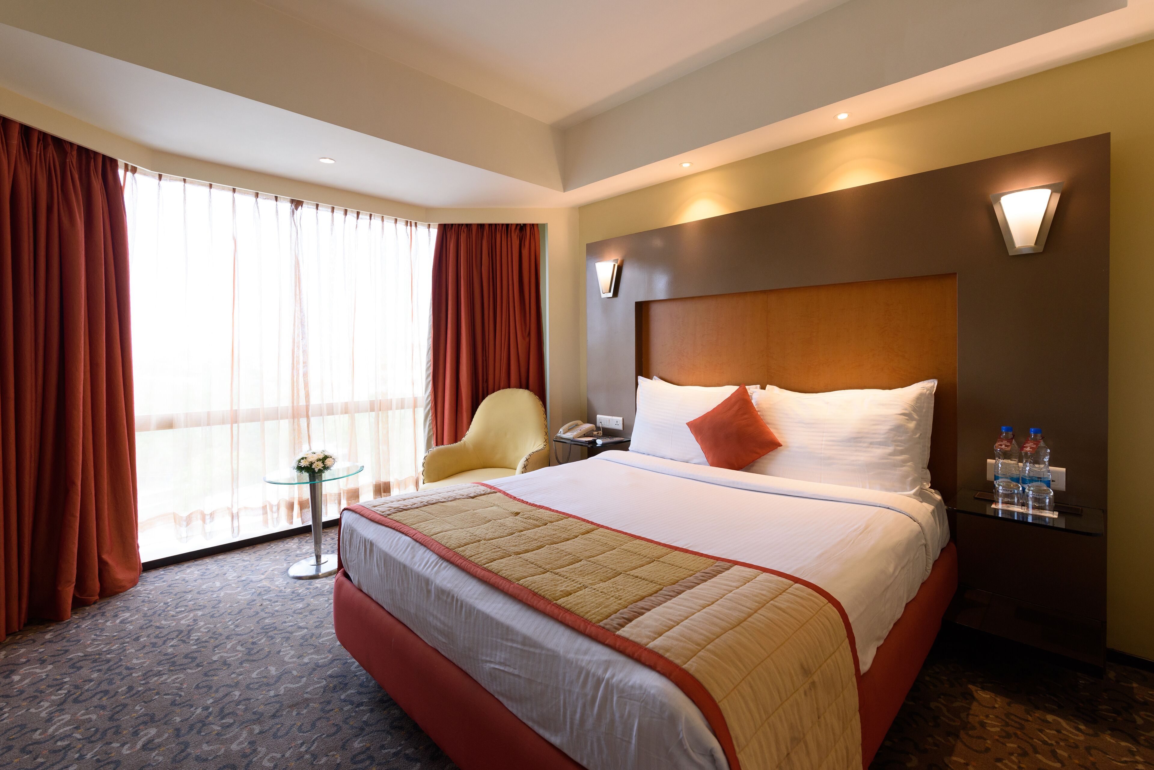 A hotel room with a large bed, a chair, a small table, and a nightstand with water bottles.