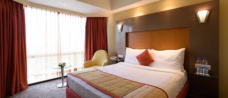 A hotel room with a large bed, a chair, a small table, and a nightstand with water bottles.