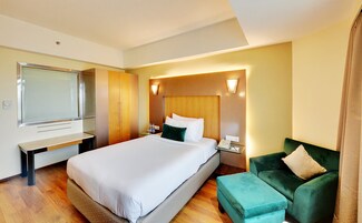 A hotel room with a large bed, a desk, a wardrobe, a green armchair, and a wall-mounted TV.
