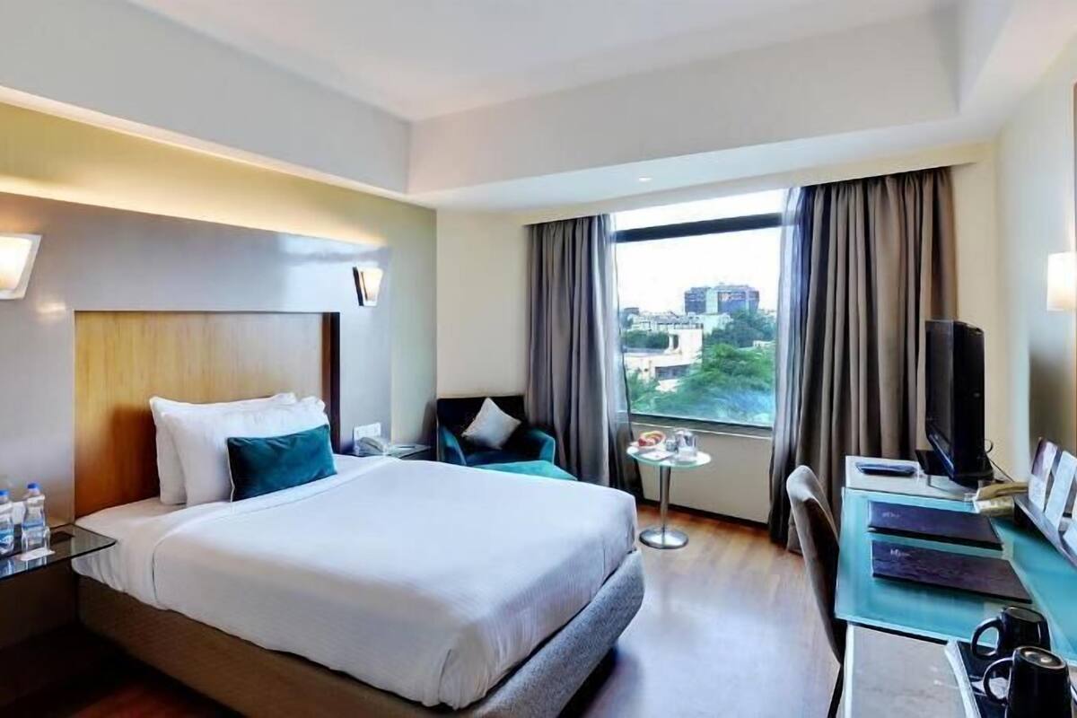 A hotel room with a bed, bedside table, desk, chair, and a view of the city.