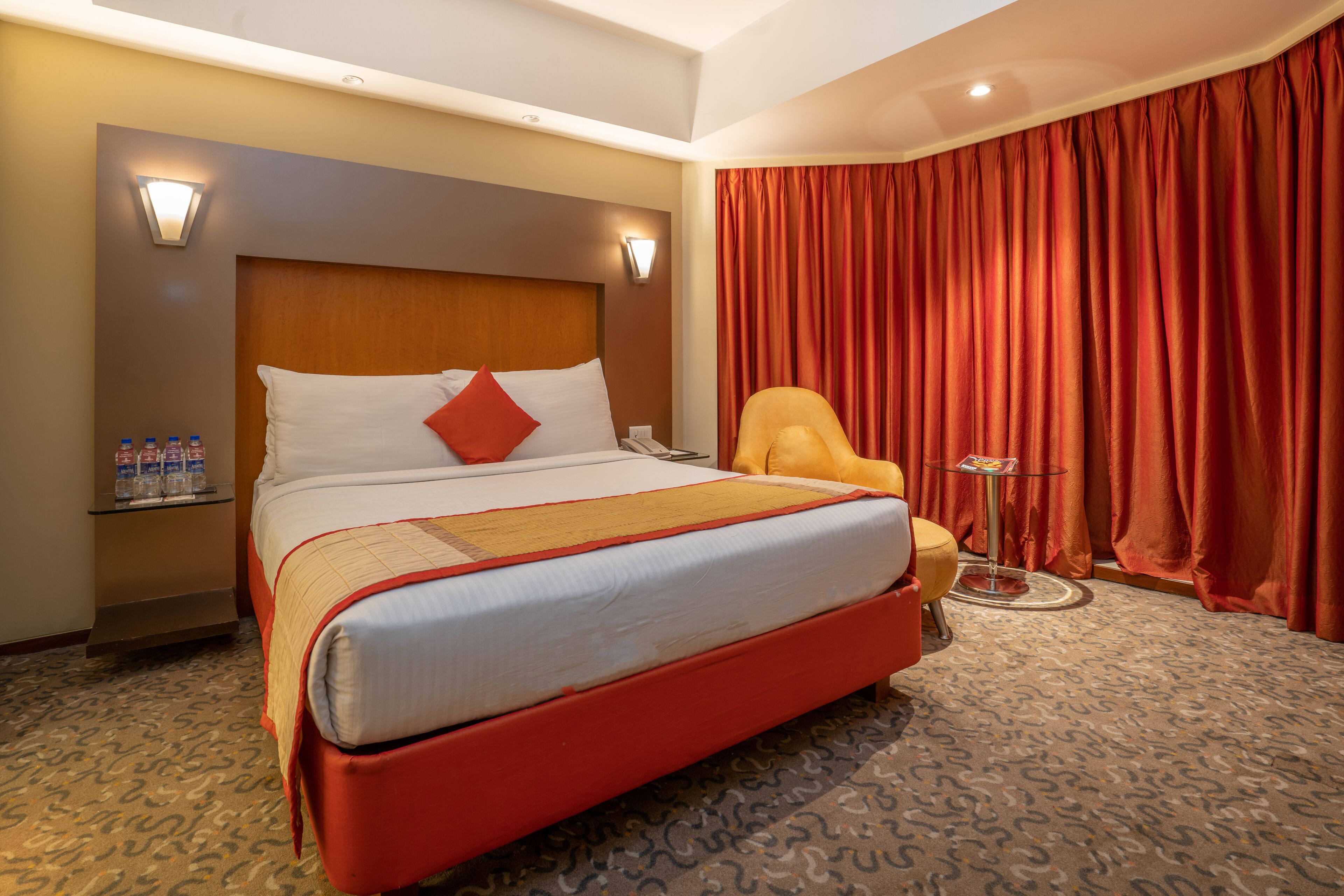 Ein Hotelzimmer mit einem großen Bett, einem Nachttisch mit Wasserflaschen, einem Sessel und einer Lampe.