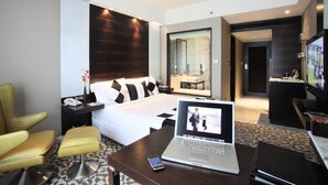 A modern hotel room with a bed, a laptop on a desk, a TV, and a seating area with a chair.