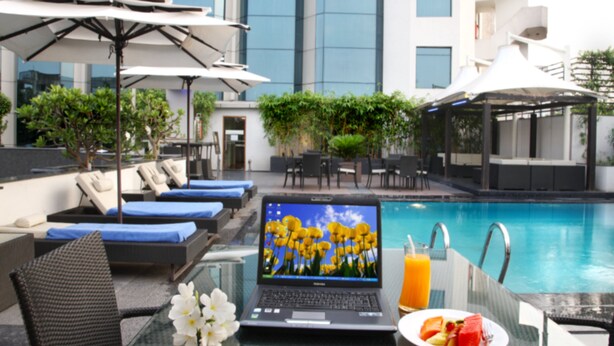 A laptop on a glass table with a plate of fruit and a glass of orange juice, overlooking a pool with lounge chairs and umbrellas.