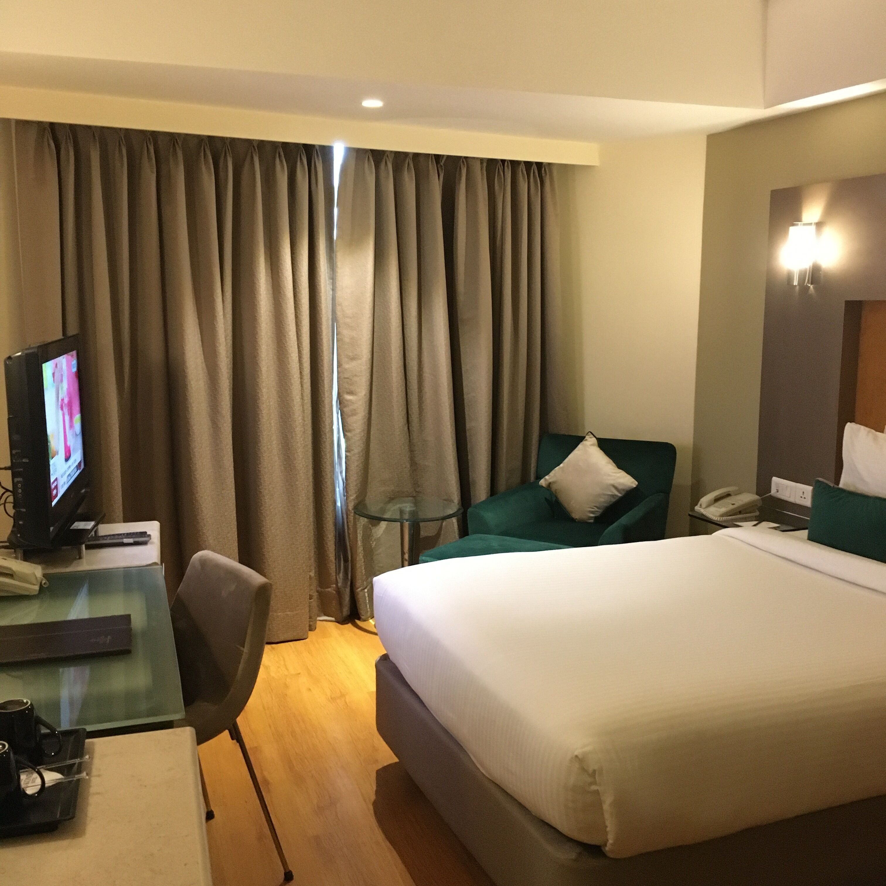 A hotel room with a bed, a desk with a television, a chair, a phone, and a window with curtains.