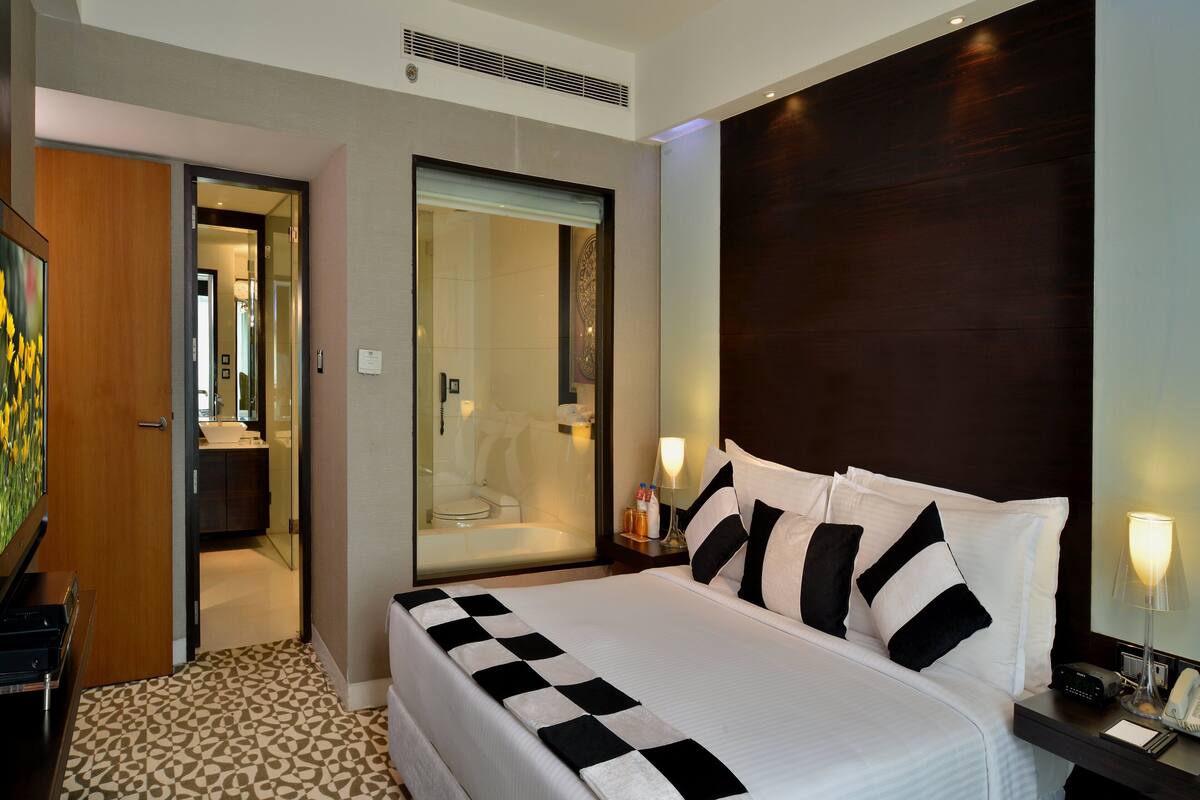 A hotel room with a large bed, a bedside table with a lamp, a bathroom with a shower, and a wooden door.