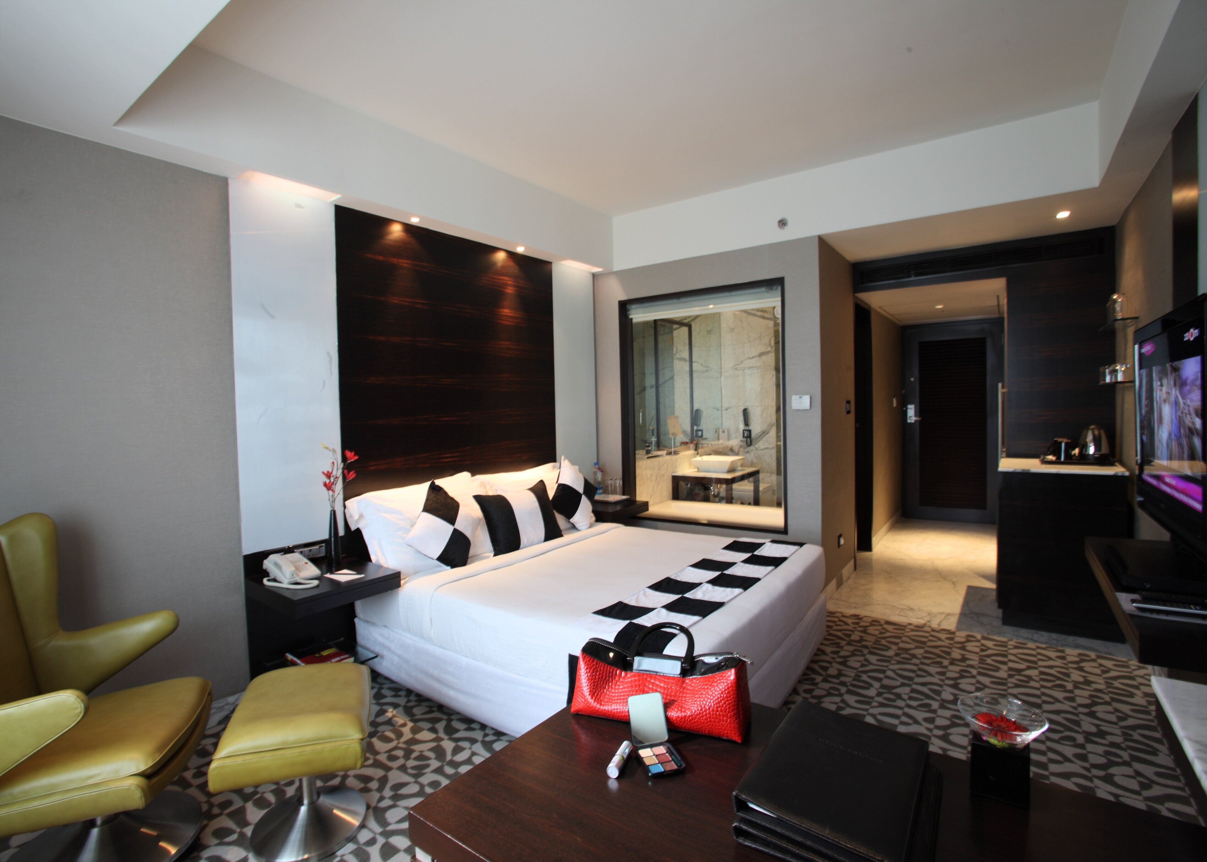 A modern hotel room with a large bed, a seating area with a chair and ottoman, a TV, and a bathroom visible through a glass partition.