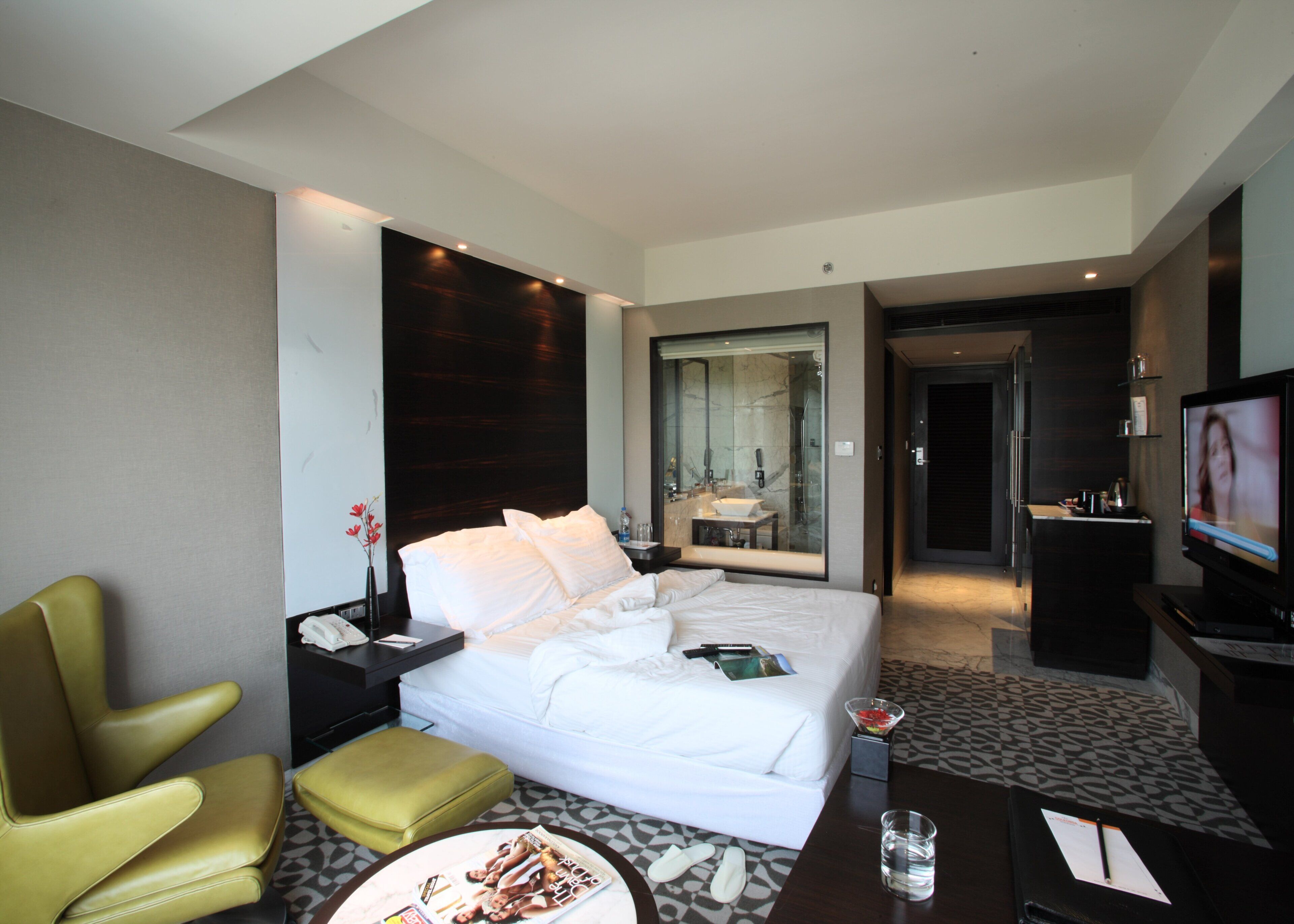 Habitación de hotel con cama, televisor, escritorio, silla y baño visible a través de una puerta de vidrio.