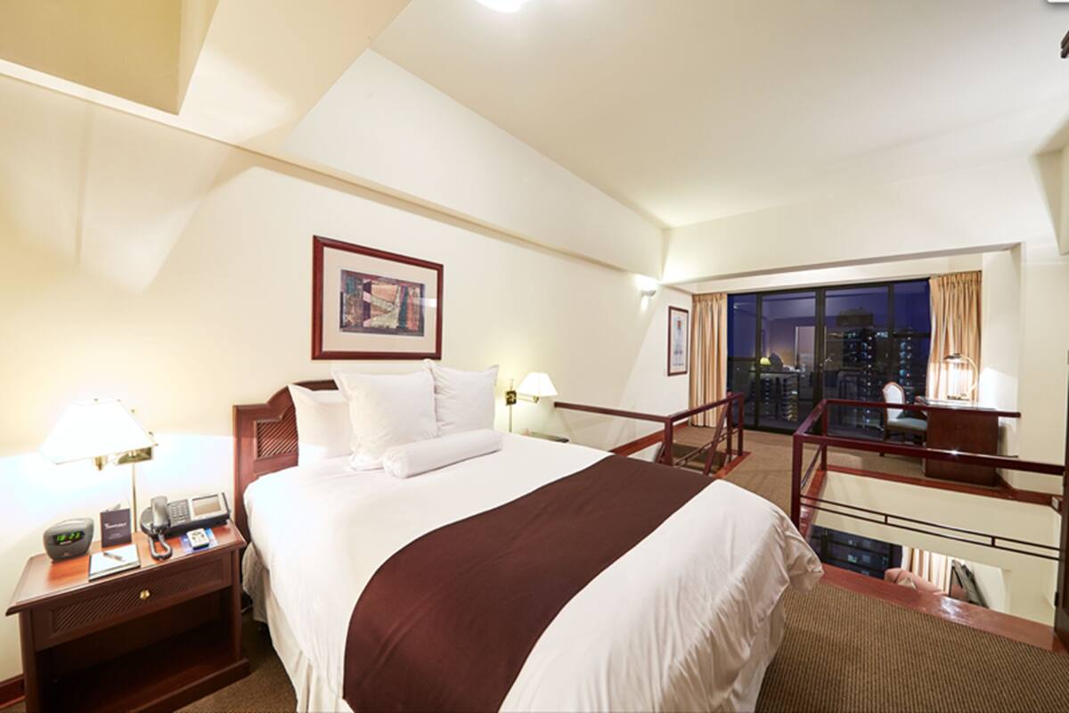 Habitación de hotel con una cama grande, un escritorio con teléfono y lámpara, una mesita de noche y vistas a la ciudad a través de un ventanal amplio.