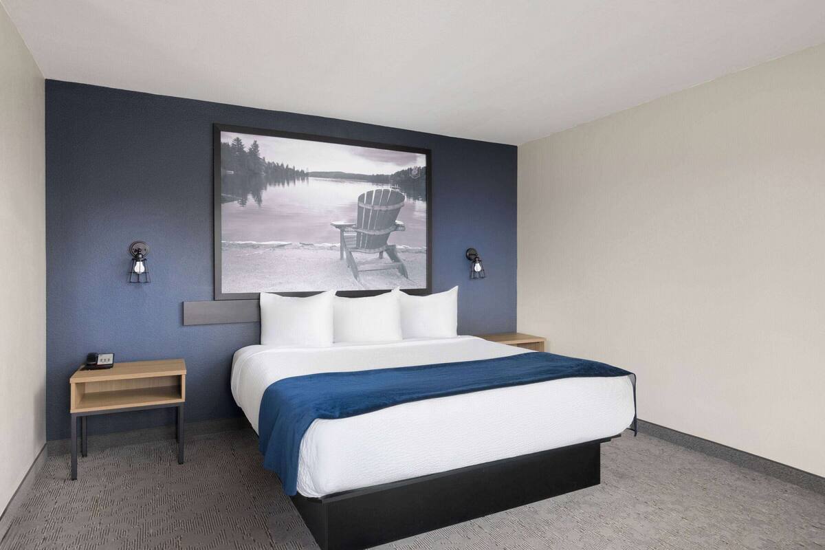 Une chambre d’hôtel moderne avec un grand lit, une tête de lit ornée d’une image représentant un paysage au bord d’un lac, deux tables de chevet avec des lampes et une petite table de nuit en bois.