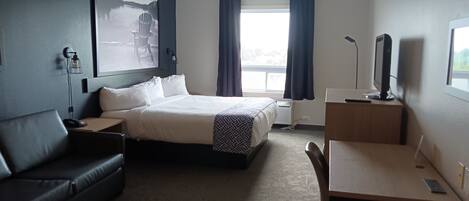 Habitación de hotel con cama, escritorio, silla, sofá, ventana grande con cortinas y televisor montado en la pared.
