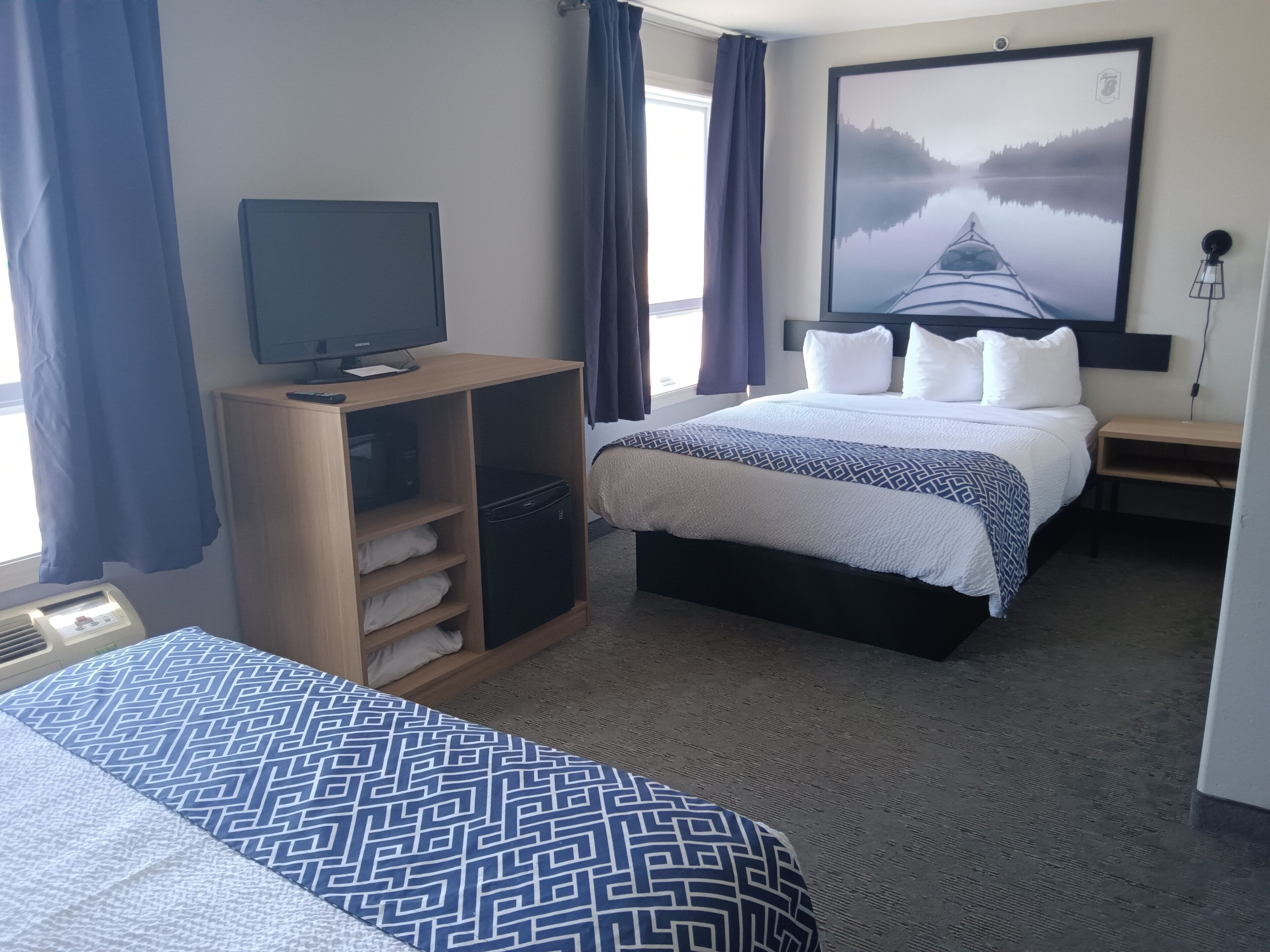A hotel room with a bed, a TV on a wooden cabinet, a large painting on the wall, and a window with curtains.