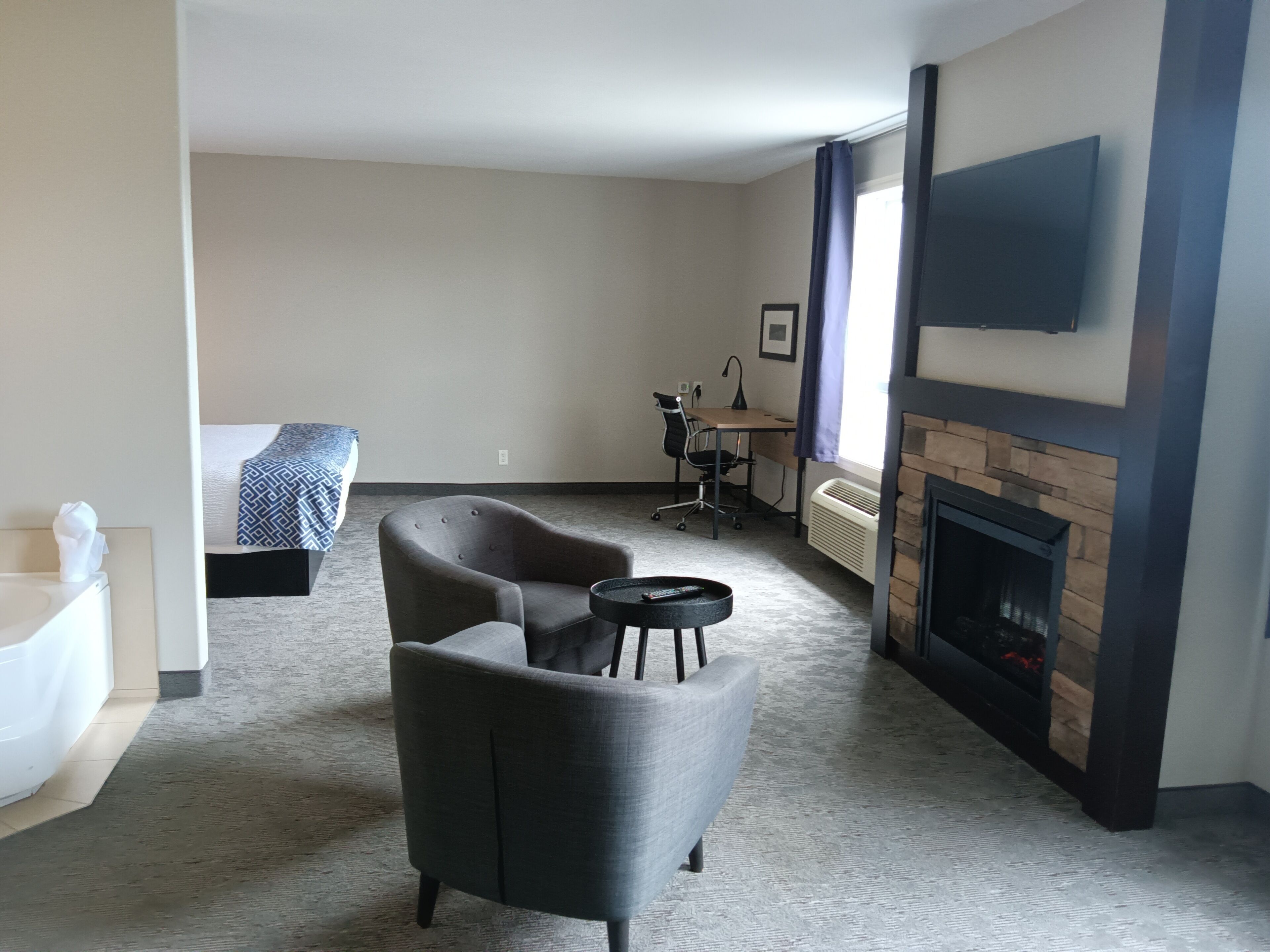 Una habitación de hotel con una cama, un sillón gris, una mesita redonda pequeña, un escritorio con una silla, un televisor de pantalla plana en la pared y una chimenea.