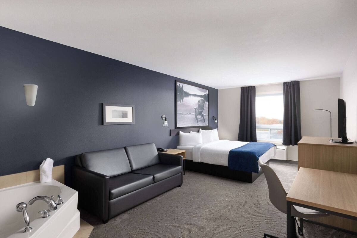 Une chambre d’hôtel avec un mur d’accent bleu foncé, un canapé en cuir noir, un lit recouvert d’une couverture bleue, un bureau en bois, une baignoire blanche et une grande fenêtre avec des rideaux.